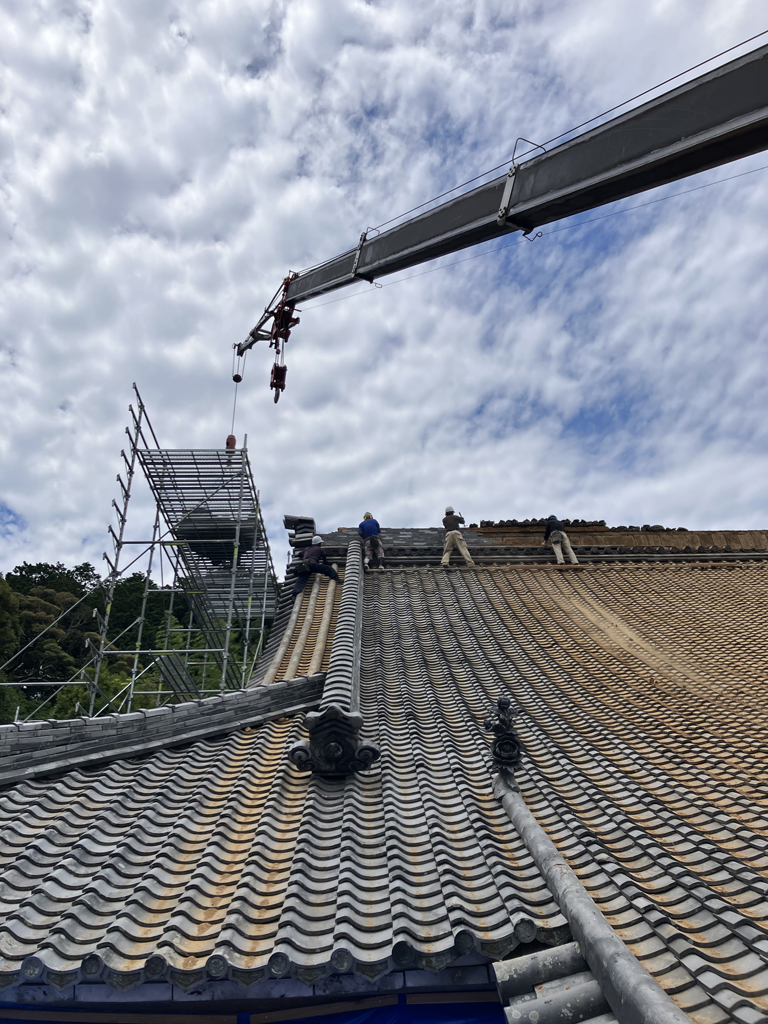 屋根リフォーム中の画像。瓦屋根の上で職人が作業している。クレーンで荷物を持ち上げている。